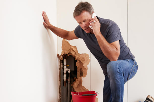 A man in bluejeans and gray t-shirt, speaking on his cellphone, with a hole in the wall behind him exposing pipes