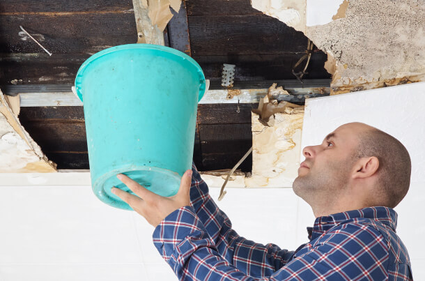 A man holding an aqua blue bucket up to a large hole in the ceiling, the surrounding ceiling looks water damaged